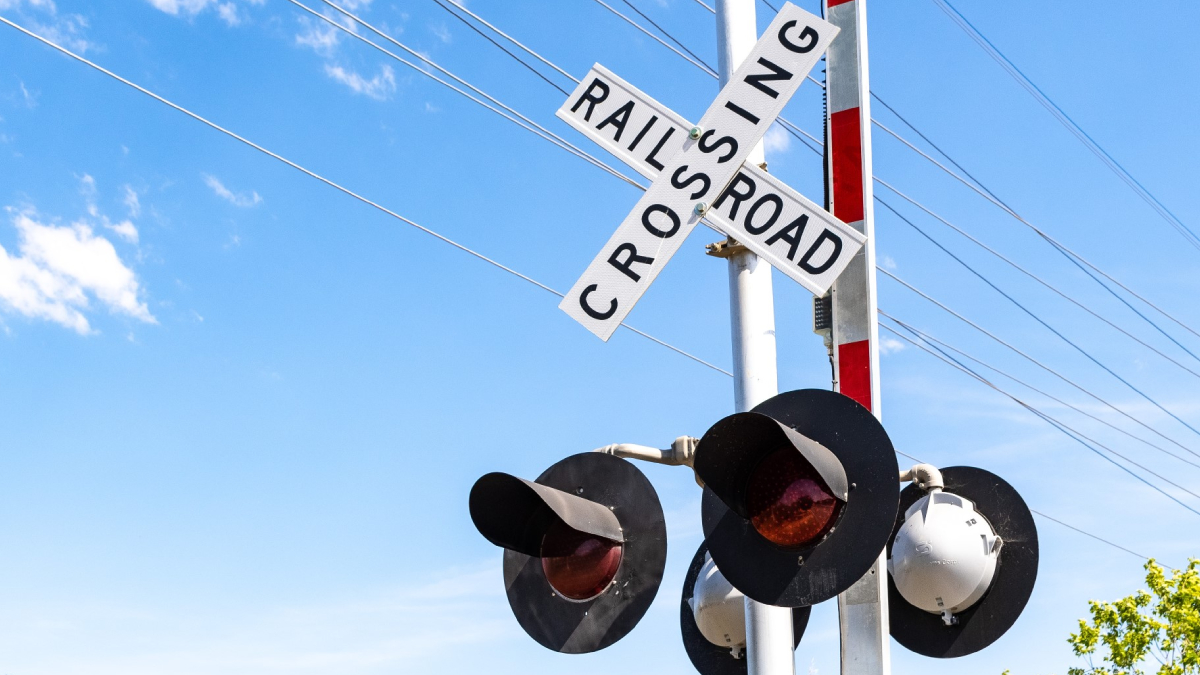 Railroad crossing sign reading 'RAILROAD CROSSING' with red warning lights on a metal pole against a blue sky and power lines behind it.