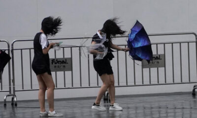 Three young performers in black outfits behind metal crowd barriers, hair blown by the wind as they move with umbrellas outstretched.