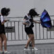 Three young performers in black outfits behind metal crowd barriers, hair blown by the wind as they move with umbrellas outstretched.