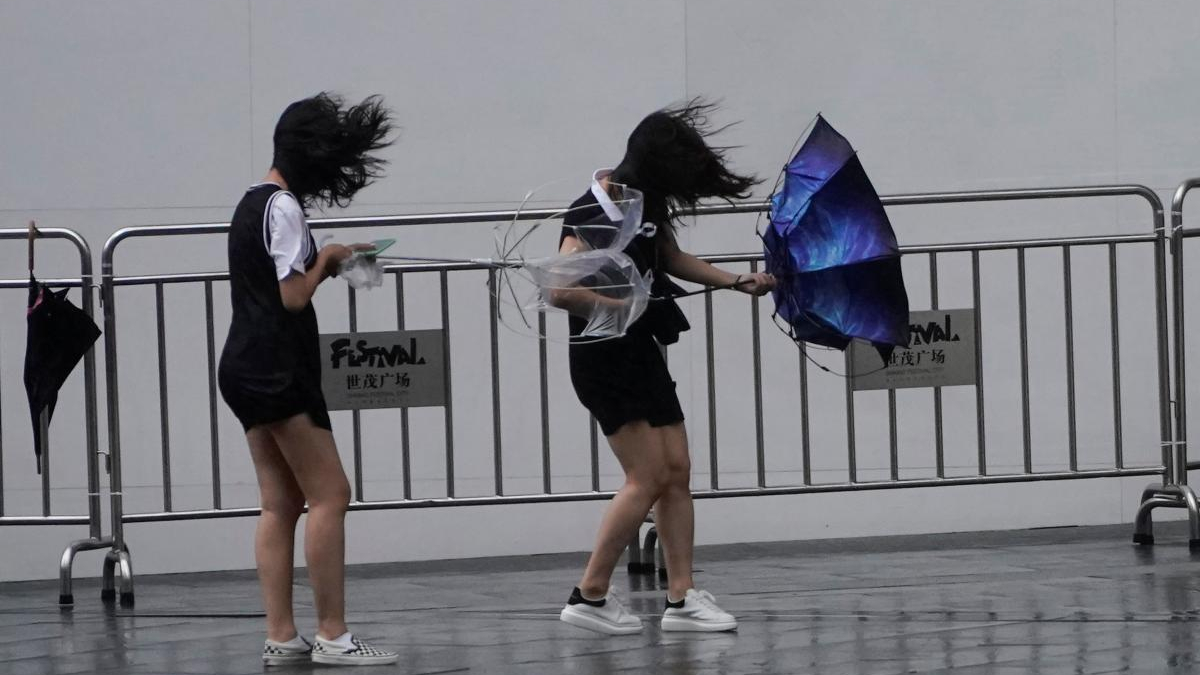 Three young performers in black outfits behind metal crowd barriers, hair blown by the wind as they move with umbrellas outstretched.