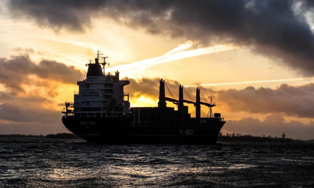 Silhouette of a large cargo ship on dark water at sunset with a bright orange and yellow sky behind it.
