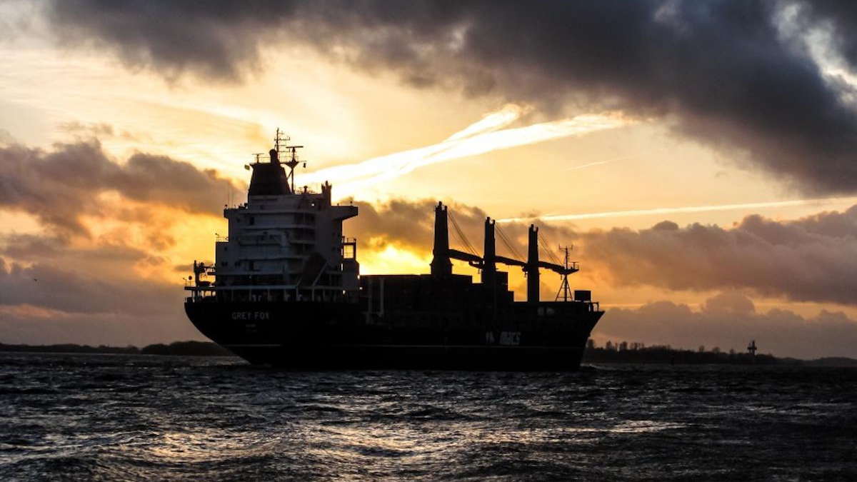 Silhouette of a large cargo ship on dark water at sunset with a bright orange and yellow sky behind it.