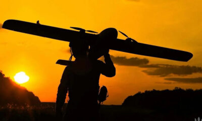 Silhouette of a person carrying a small propeller plane over their shoulder at sunset, orange sky in the background.