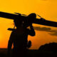 Silhouette of a person carrying a small propeller plane over their shoulder at sunset, orange sky in the background.