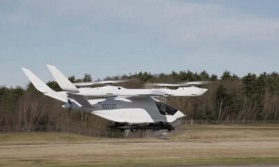 White tilt-rotor aircraft lifting off from a runway, with dual wing-mounted rotors and a compact cockpit.