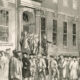 Historic crowd gathered outside a grand neoclassical building with tall columns and arched entrance.