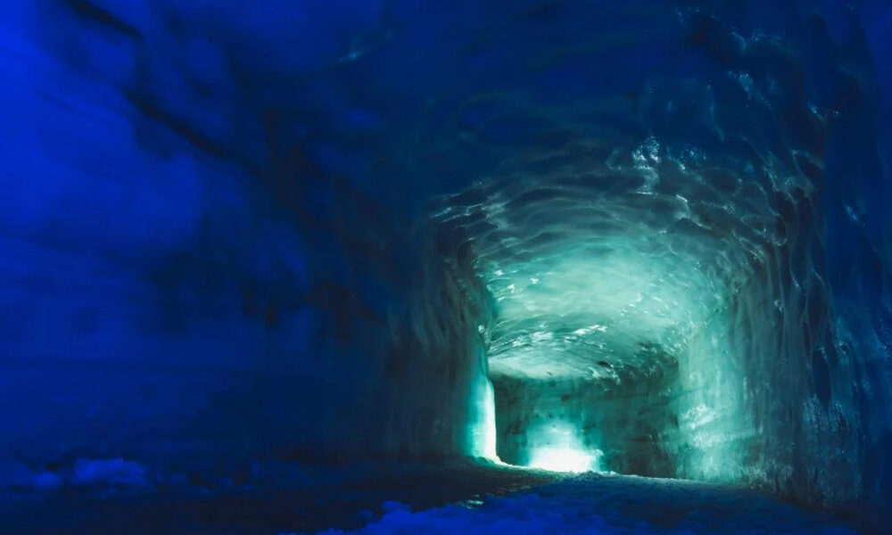 Ice cave tunnel with blue ice walls and a turquoise glow at the far end of the tunnel.