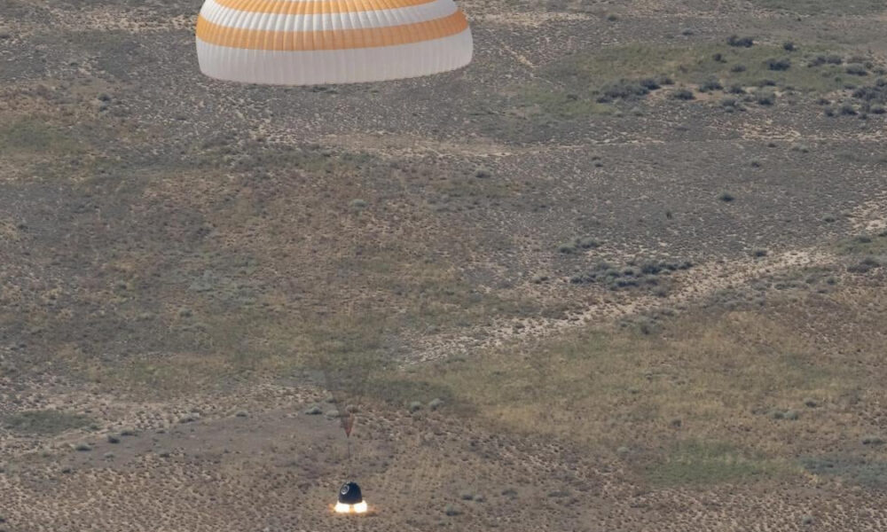 Desert landscape with a white and orange circular parachute drifting above and a bright flame on the ground below, suggesting a landing or takeoff.