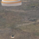 Desert landscape with a white and orange circular parachute drifting above and a bright flame on the ground below, suggesting a landing or takeoff.