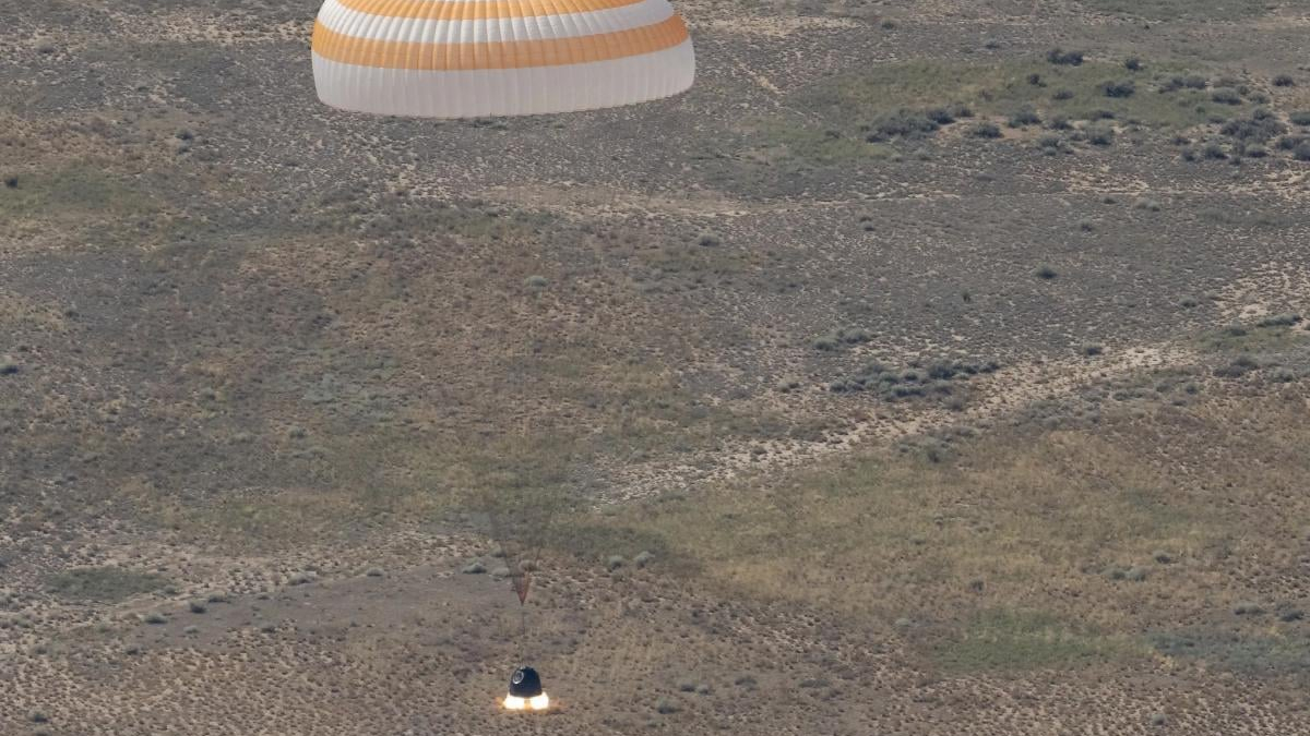 Desert landscape with a white and orange circular parachute drifting above and a bright flame on the ground below, suggesting a landing or takeoff.