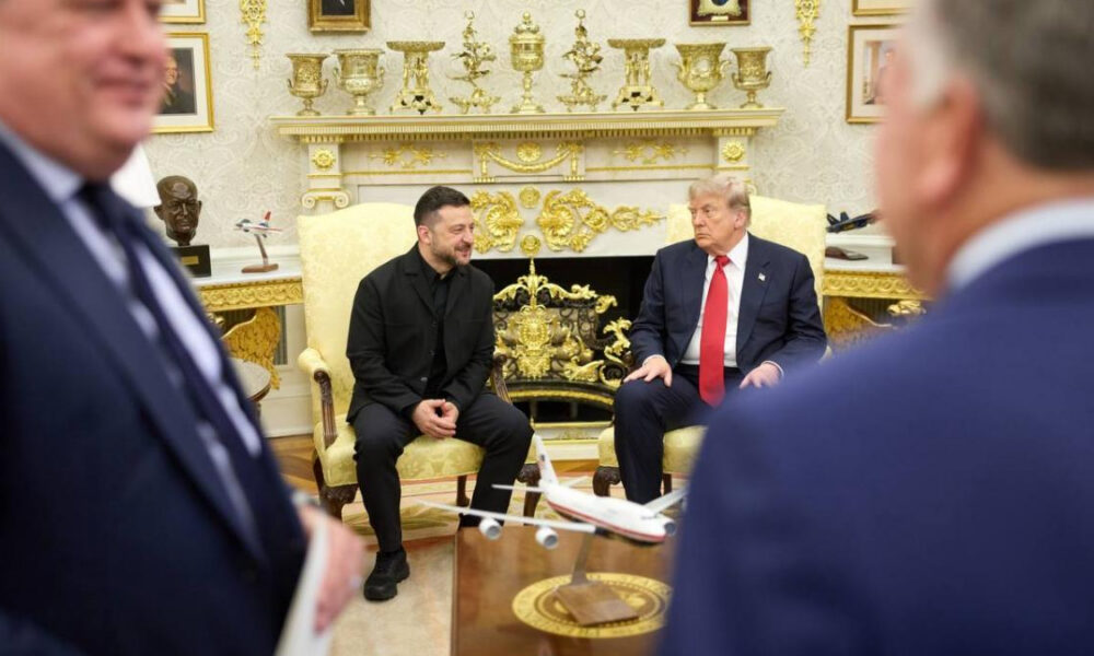 Two men converse in a gold-decorated room, seated in yellow armchairs, with a small model airplane on the table between them.