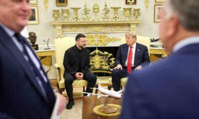 Two men in dark suits sit on gold-upholstered chairs in a lavish room; a model airplane sits on a table between them.