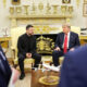 Two men in dark suits sit on gold-upholstered chairs in a lavish room; a model airplane sits on a table between them.