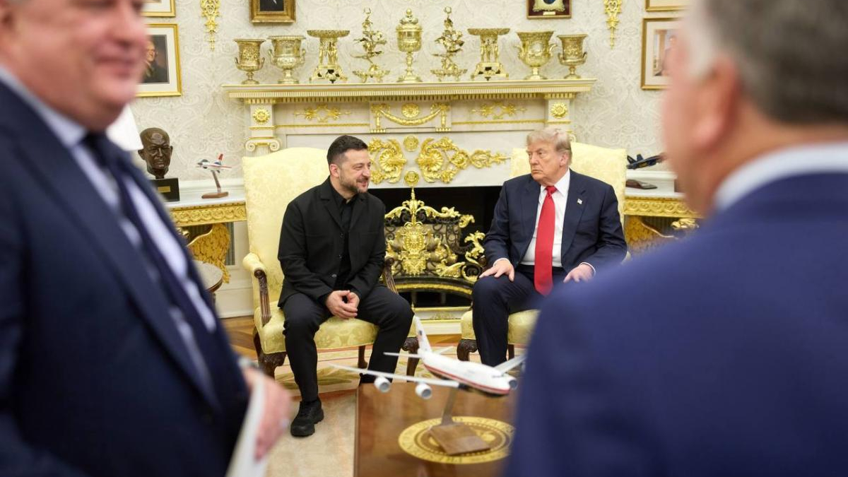 Two men in dark suits sit on gold-upholstered chairs in a lavish room; a model airplane sits on a table between them.