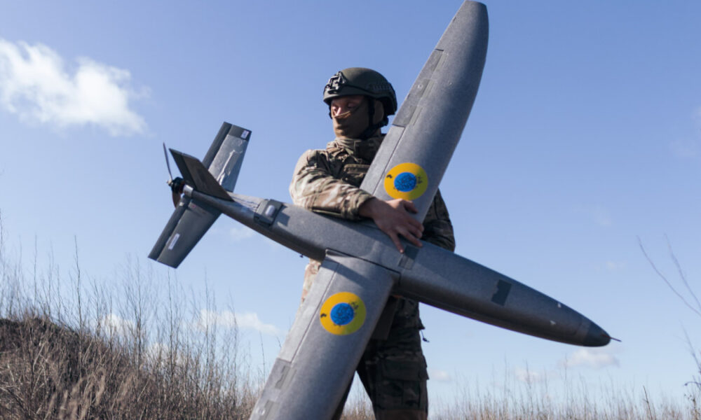 Soldier in camouflage uniform and helmet holding a large gray unmanned drone outdoors in a grassy field under a blue sky.
