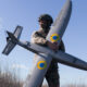 Soldier in camouflage uniform and helmet holding a large gray unmanned drone outdoors in a grassy field under a blue sky.