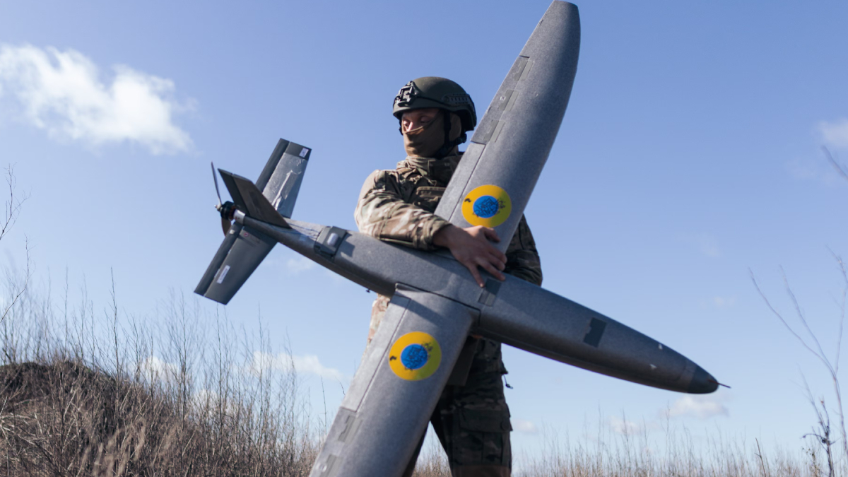 Soldier in camouflage uniform and helmet holding a large gray unmanned drone outdoors in a grassy field under a blue sky.