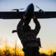 Soldier in camouflage gear lifts a small propeller airplane overhead at sunset, silhouetted against a golden sky from behind the trees.