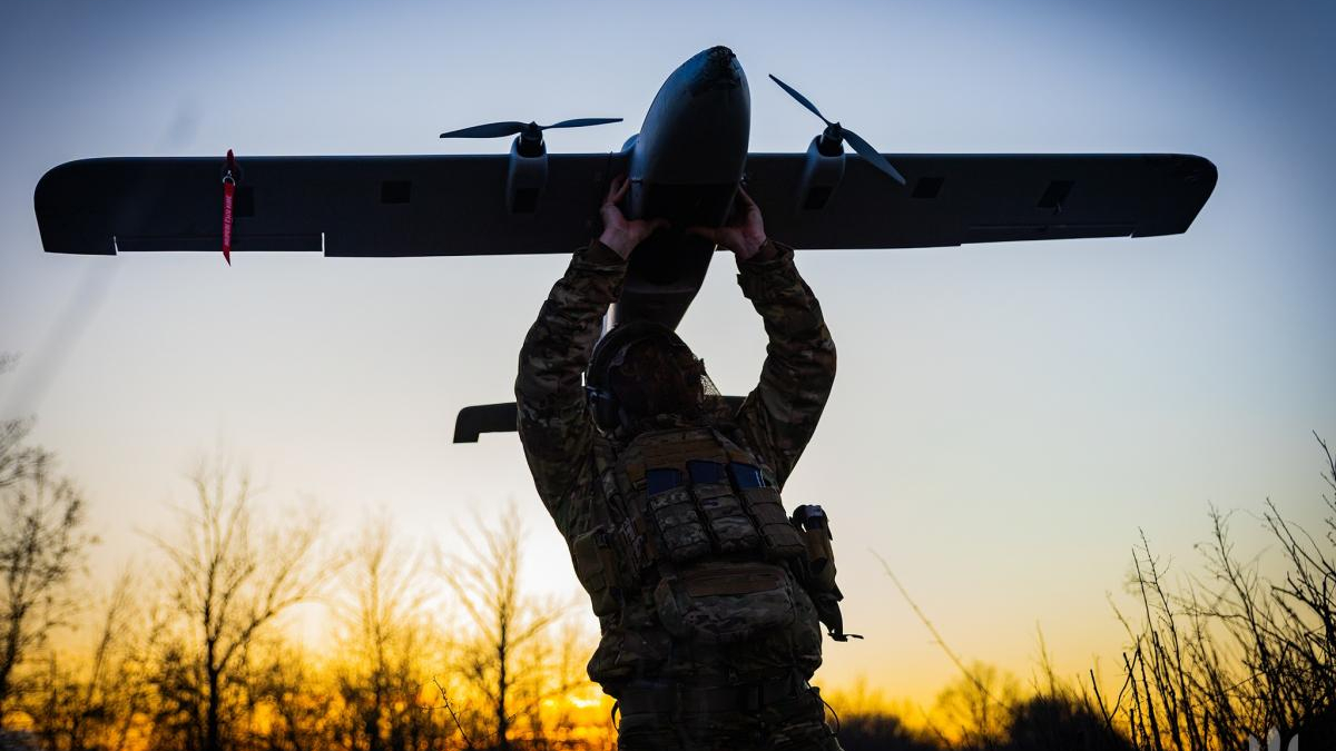 Soldier in camouflage gear lifts a small propeller airplane overhead at sunset, silhouetted against a golden sky from behind the trees.