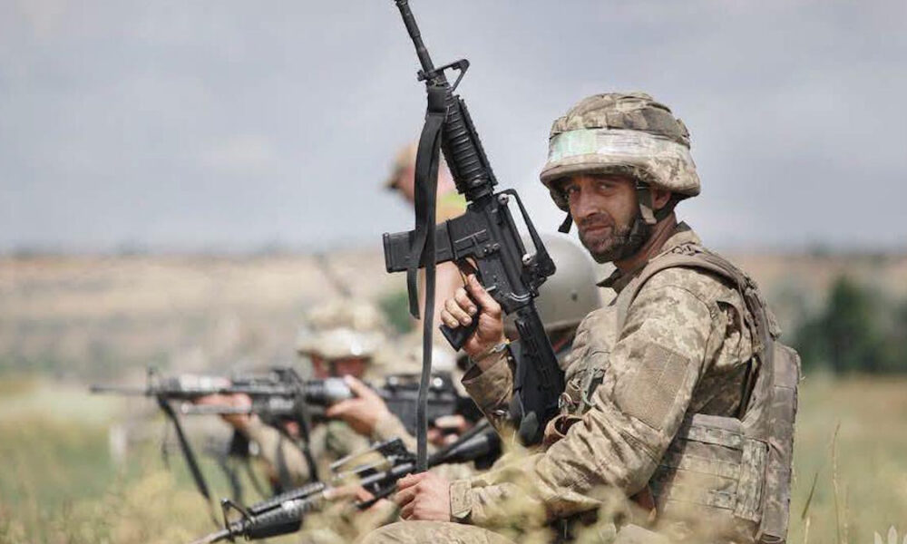 Soldier in camouflage helmet and gear aiming a rifle in a field, lined up with others behind him.