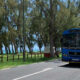 Blue tour bus driving along a coastal road with trees and the sea in the background.