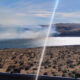 Desert shoreline with sparse shrubs, a sunlit lake, and distant brown hills under a clear blue sky.