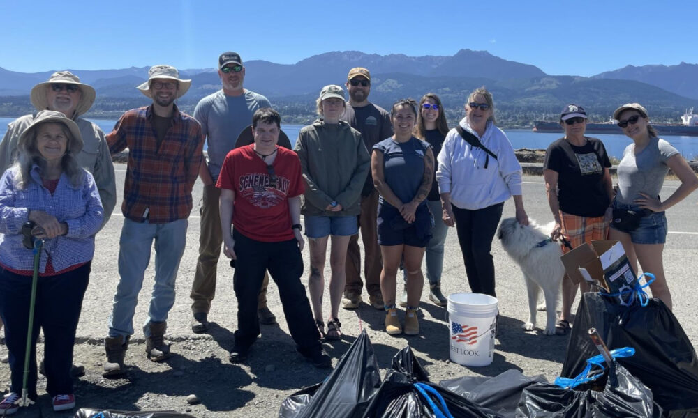 Group of volunteers posing together on a waterfront road after a shoreline cleanup, with trash bags, a white bucket with a flag, and a dog nearby, mountains in the background.