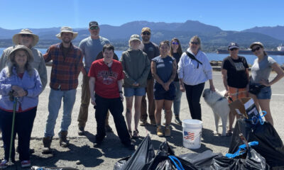 Group of volunteers posing together on a waterfront road after a shoreline cleanup, with trash bags, a white bucket with a flag, and a dog nearby, mountains in the background.
