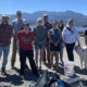 Group of volunteers posing together on a waterfront road after a shoreline cleanup, with trash bags, a white bucket with a flag, and a dog nearby, mountains in the background.