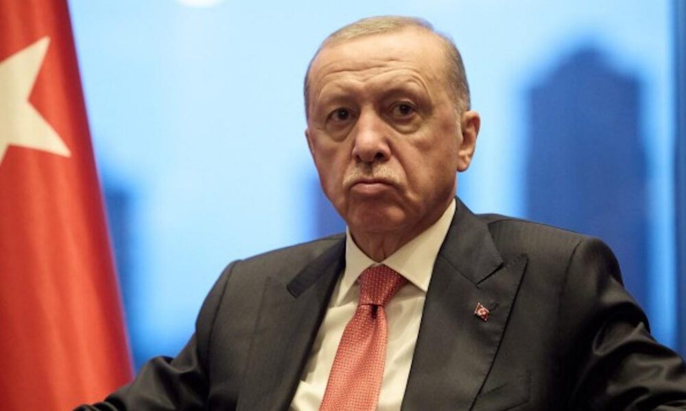 Man in a dark suit and red tie sits at a formal event, with a Turkish flag visible on the left.