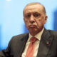Man in a dark suit and red tie sits at a formal event, with a Turkish flag visible on the left.