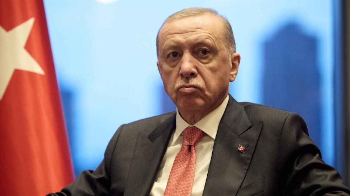 Man in a dark suit and red tie sits at a formal event, with a Turkish flag visible on the left.