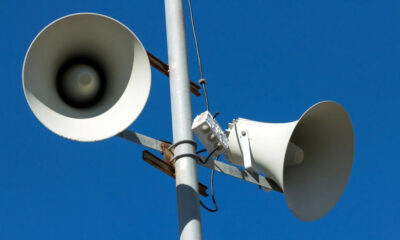 Public address speakers mounted on a metal pole against a clear blue sky.
