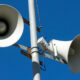 Public address speakers mounted on a metal pole against a clear blue sky.