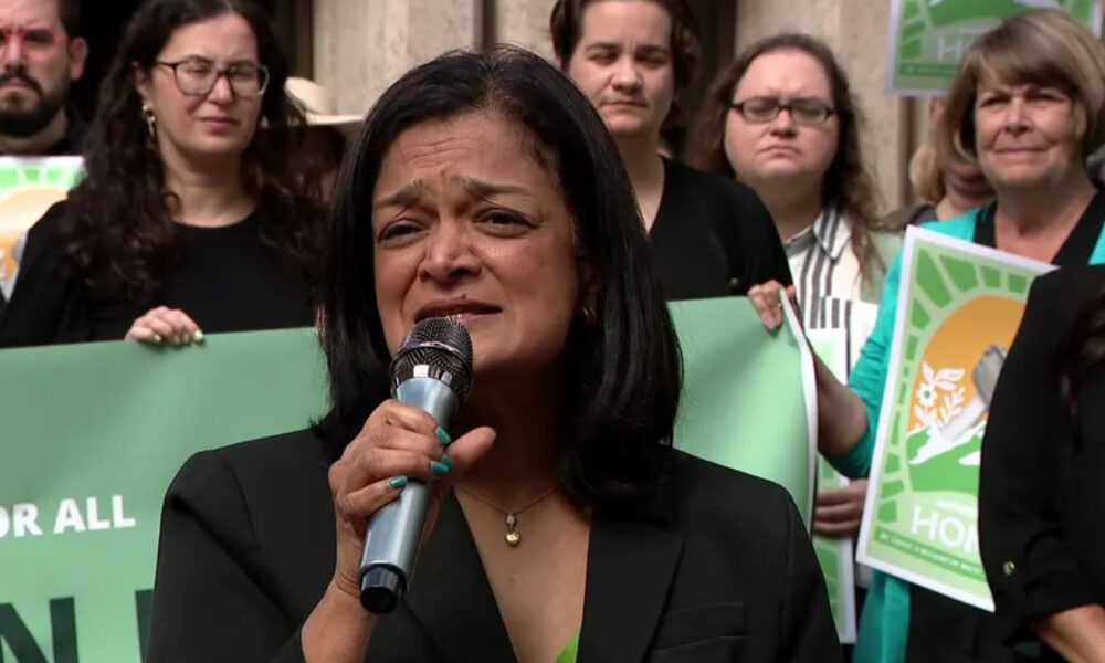 Woman in a black blazer speaks into a microphone at a rally, with supporters holding green protest signs behind her.