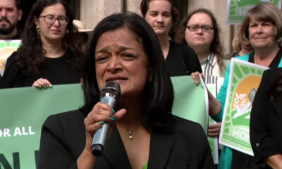 Woman in a black blazer speaks into a microphone at a rally, with supporters holding green protest signs behind her.