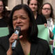 Woman in a black blazer speaks into a microphone at a rally, with supporters holding green protest signs behind her.