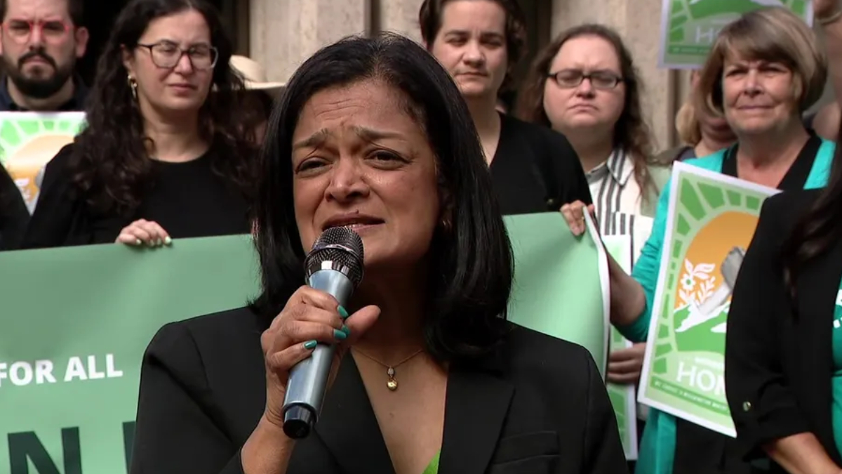 Woman in a black blazer speaks into a microphone at a rally, with supporters holding green protest signs behind her.