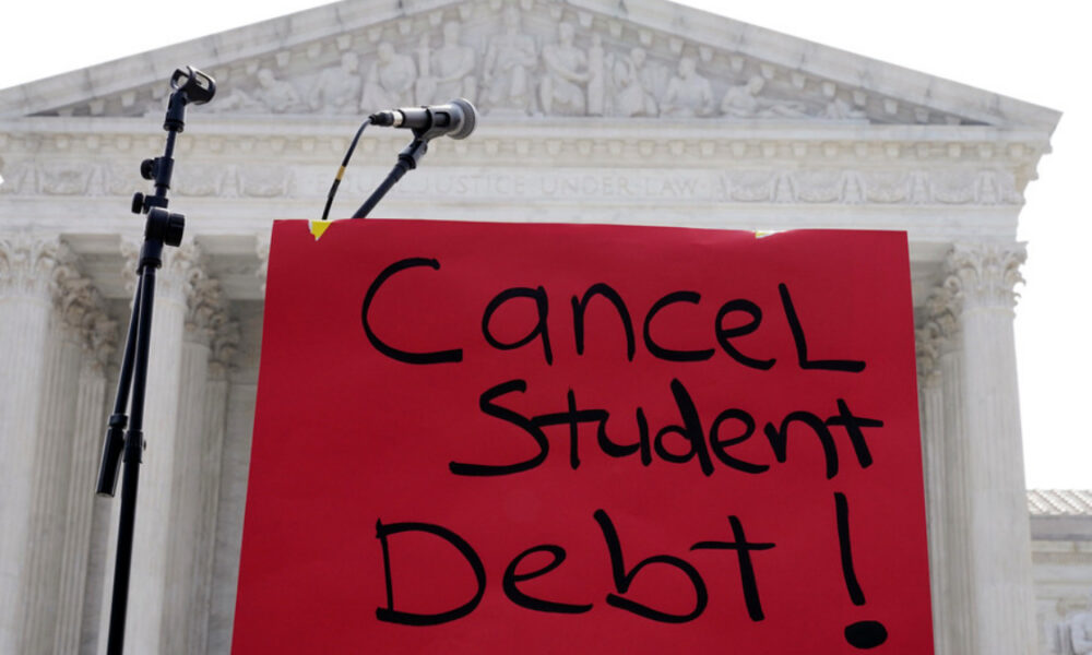 Red protest sign reading 'Cancel Student Debt!' with microphones in front of a classical courthouse backdrop.