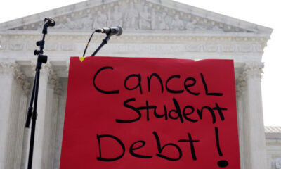 Red protest sign reading 'Cancel Student Debt!' with microphones in front of a classical courthouse backdrop.