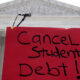 Red protest sign reading 'Cancel Student Debt!' with microphones in front of a classical courthouse backdrop.