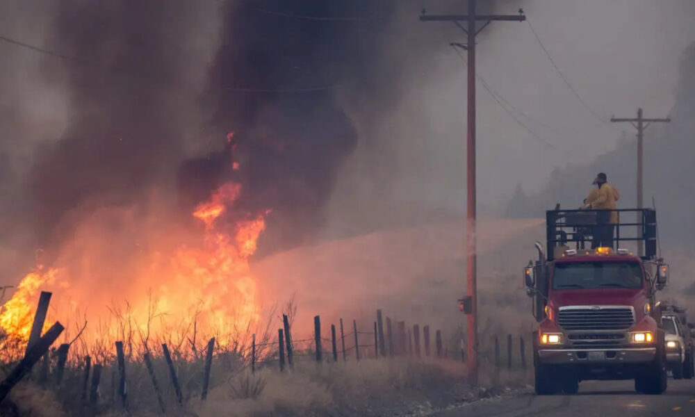 Fire truck with a raised platform containing a firefighter spraying water at a grass fire along a rural road as flames and thick smoke rise nearby.