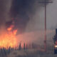 Fire truck with a raised platform containing a firefighter spraying water at a grass fire along a rural road as flames and thick smoke rise nearby.