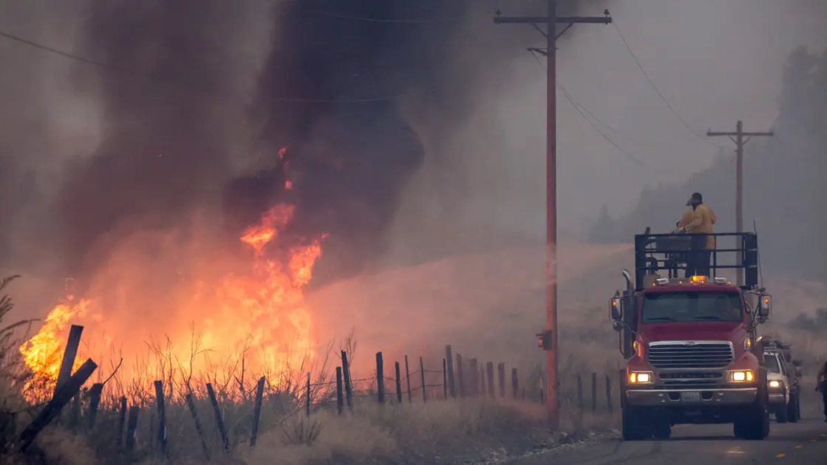 Fire truck with a raised platform containing a firefighter spraying water at a grass fire along a rural road as flames and thick smoke rise nearby.