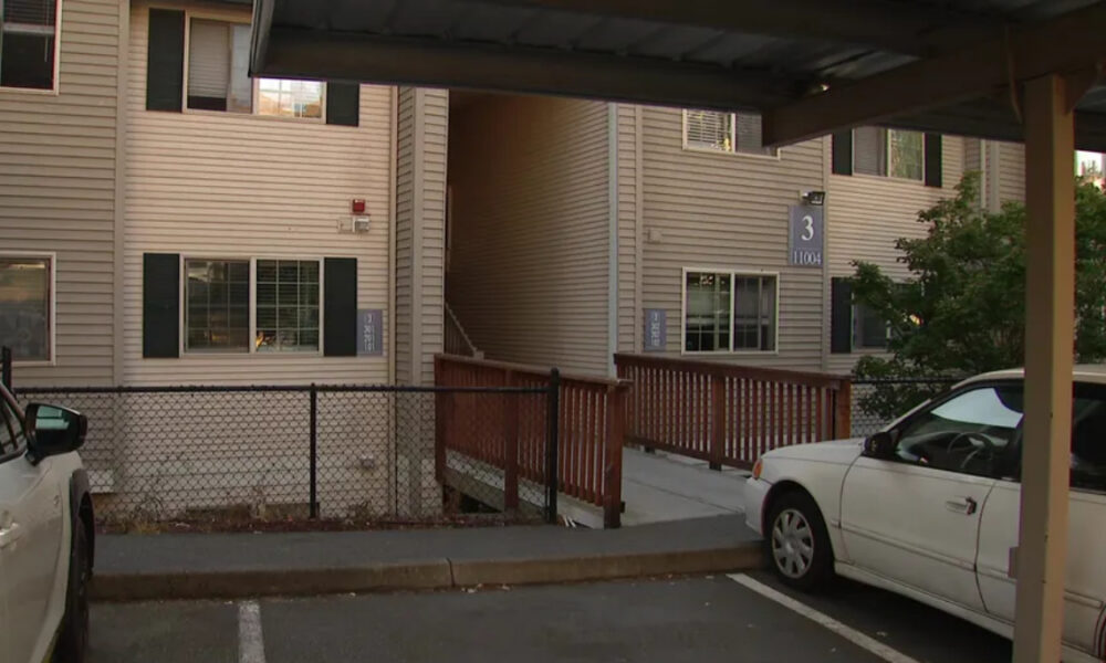 Two-story beige apartment building with a wooden ramp leading to stairs, a chain-link fence, and parked cars in the foreground.