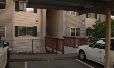 Two-story beige apartment building with a wooden ramp leading to stairs, a chain-link fence, and parked cars in the foreground.