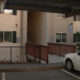 Two-story beige apartment building with a wooden ramp leading to stairs, a chain-link fence, and parked cars in the foreground.