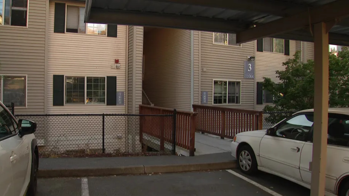 Two-story beige apartment building with a wooden ramp leading to stairs, a chain-link fence, and parked cars in the foreground.
