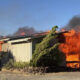 A single-story shed engulfed in flames with thick black smoke rising above it.
