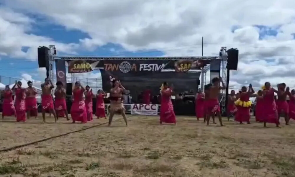Group of dancers in pink skirts performing on a sandy festival ground in front of a stage with a banner overhead reading Festival and related logos from the event stage area.